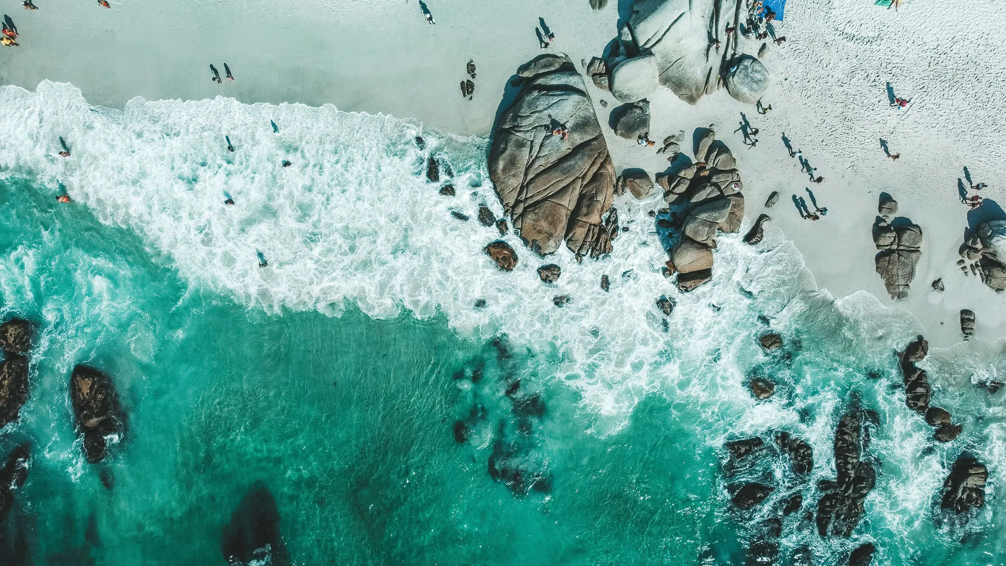 Cape Town coastline from directly above, turquoise surf breaking over granite boulders onto white sand