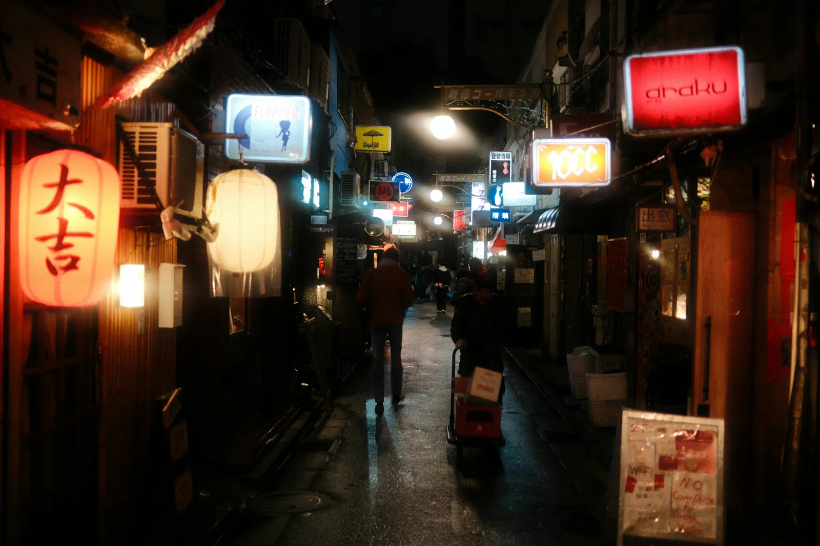 A lantern-lit alley in Golden Gai at night, bar signs crowding both walls above wet pavement