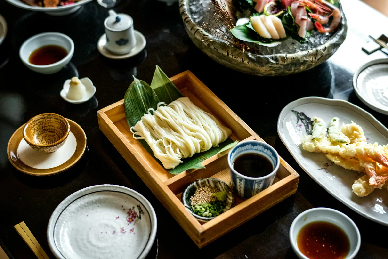 A set meal on a dark table: chilled noodles in a wooden box, tempura, sashimi on ice, and dipping bowls