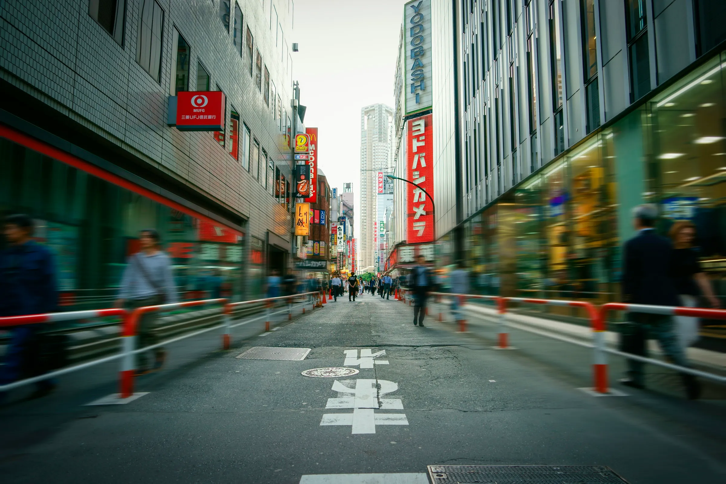 A narrow Shinjuku shopping street at dusk, signs stacked up both sides and commuters blurred past the barriers