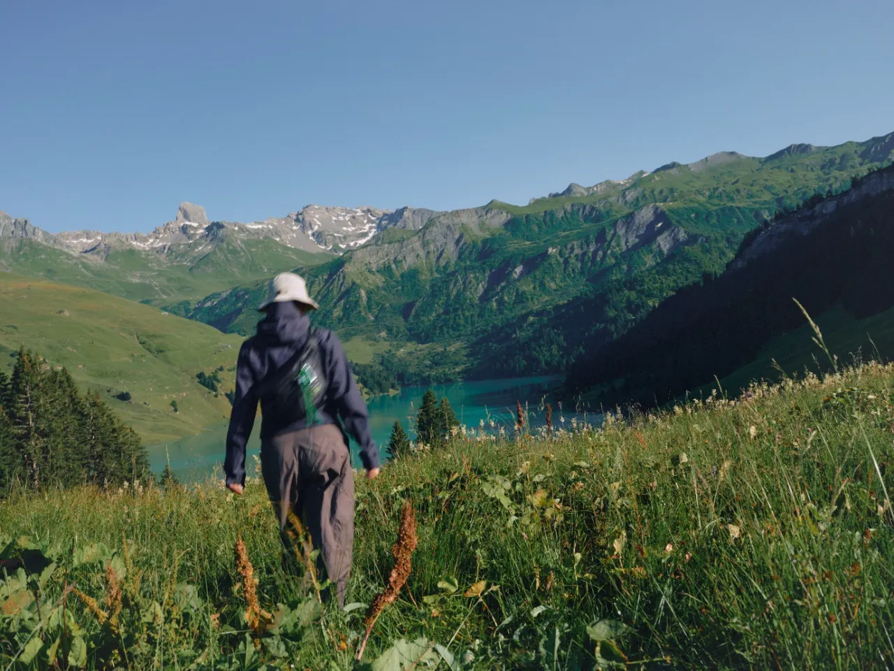 A hiker walking through a tall-grass meadow toward a turquoise alpine lake