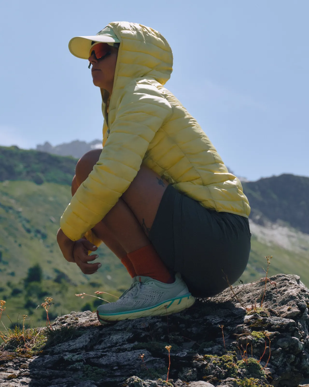 A hiker in a yellow puffer jacket crouched on a rock above a green alpine valley
